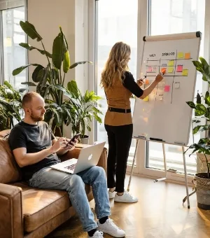UX strategist mapping sprint tasks on flip-chart while developer reviews code on phone and laptop, Studio Ubique office