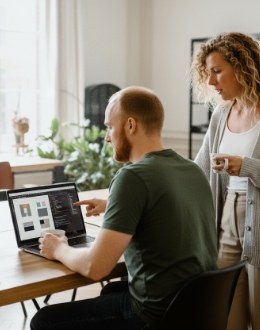 Collega’s bekijken samen een ontwerp op een laptop, één wijst naar het scherm terwijl de ander meekijkt met koffie.