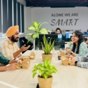 Teamoverleg aan een tafel met laptops, planten en een slogan op de muur op de achtergrond.