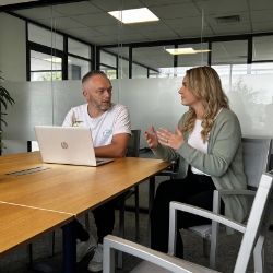 Twee collega’s overleggen in een vergaderruimte met glaswand, met laptop op tafel.