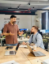 Collega licht iets toe terwijl een ander luistert aan een bureau met twee laptops in een kantoor met planten.