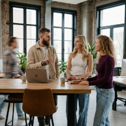 Three Studio Ubique colleagues in a standing discussion around a high wooden table with MacBook, fourth colleague walking past with motion blur