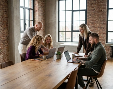 Vijf Studio Ubique collega's werken samen rond een lange houten tafel met MacBooks in het Zwolse industriële kantoor, schuin van bovenaf
