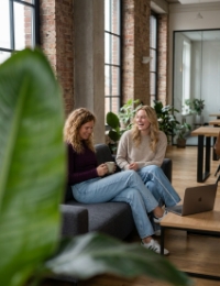 Two colleagues taking a break on a sofa in a Zwolle industrial office, plant in blurred foreground, brick wall and daylight