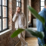 Collega in telefoongesprek leunend bij een hoog industrieel raam, bakstenen muur en plant wazig in voorgrond