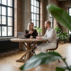Two colleagues in a project scope conversation at a round wooden table in the Zwolle industrial office, coffee and MacBook between them