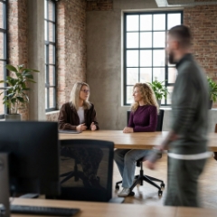 Twee Studio Ubique collega's in gesprek aan een houten bureau in het Zwolse industriële kantoor, collega loopt voorbij met bewegingsblur