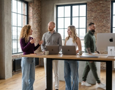 Three Studio Ubique colleagues in standing discussion around a high wooden table with MacBooks, fourth colleague walking past with motion blur