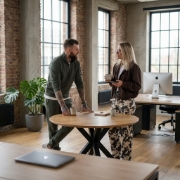 Two colleagues meeting at a round wooden table in an industrial Zwolle office, overhead angle, MacBook and daylight from tall windows