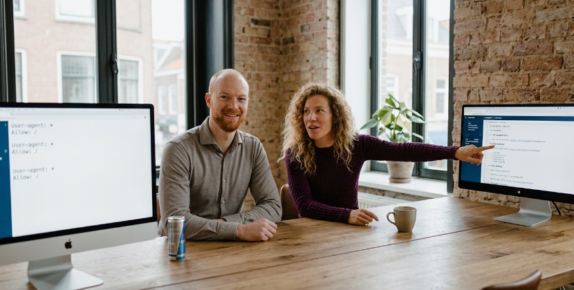 Twee collega's aan een houten bureau in Zwolle vergelijken robots.txt en llms.txt op twee schermen
