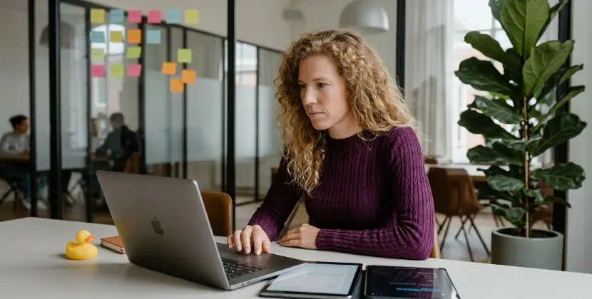 Designer in a glass meeting room in Zwolle comparing a layout on a laptop and tablet side by side, sticky notes behind
