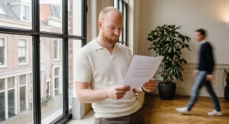 Person by a tall office window, reading a printed page in the daylight