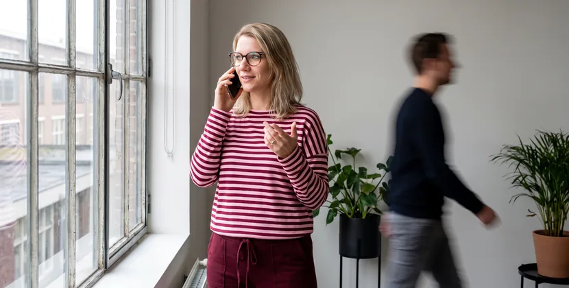 Woman by an office window on a phone call, checking a printed sheet